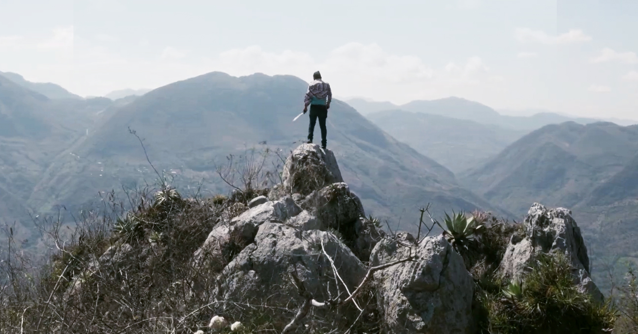 Un hombre de espaldas, con un machete en la mano, está sobre unas rocas mirando un paisaje de montañas.