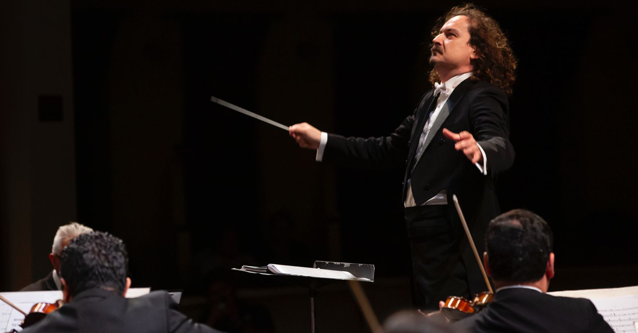 Imagen del director de orquesta Gil de Gálvez durante un concierto y dos músicos, violinistas, de espaldas.