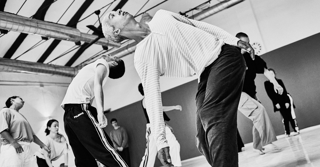 Varios bailarines y bailarinas jóvenes, vestidos de manera deportiva, se mueven en una sala. Es una imagen en blanco y negro.