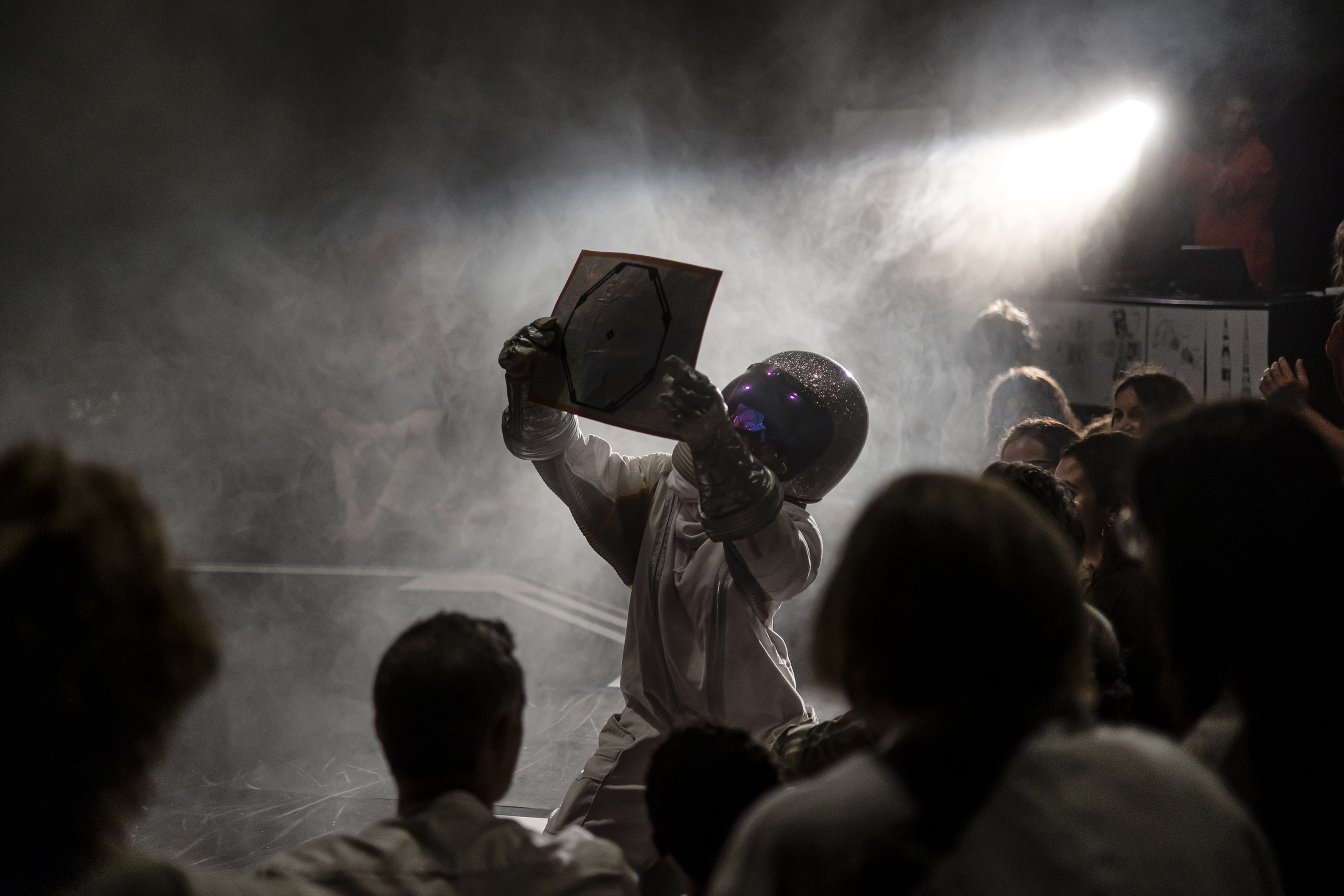 La imagen muestra a una persona vestida con escafandra y casco de cosmonauta, arrodillada en medio de un escenario circular y lleno de humo, ...