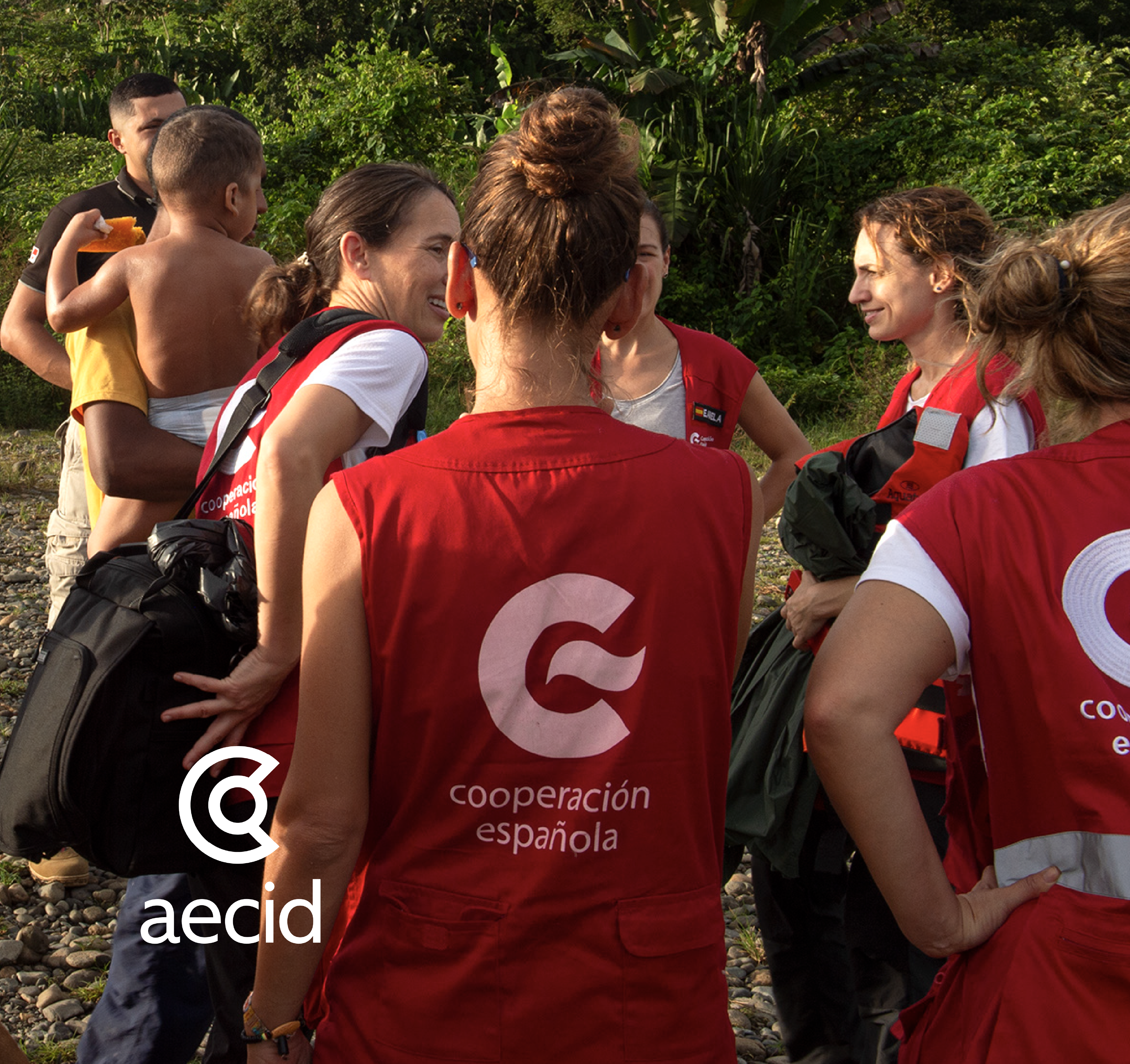 Foto de un grupo de mujeres jóvenes vestidas con unos chalecos rojos que tienen en la espalda y el logotipo de la C y las palabras Cooperación Española. Junto a ella hay dos muchachos jóvenes y uno de ellos carga un niño pequeño. En la parte inferior izquierda aparece en color blanco el logo de la AECID que es una C de líneas gruesas que en el medio se cierra en un círculo.