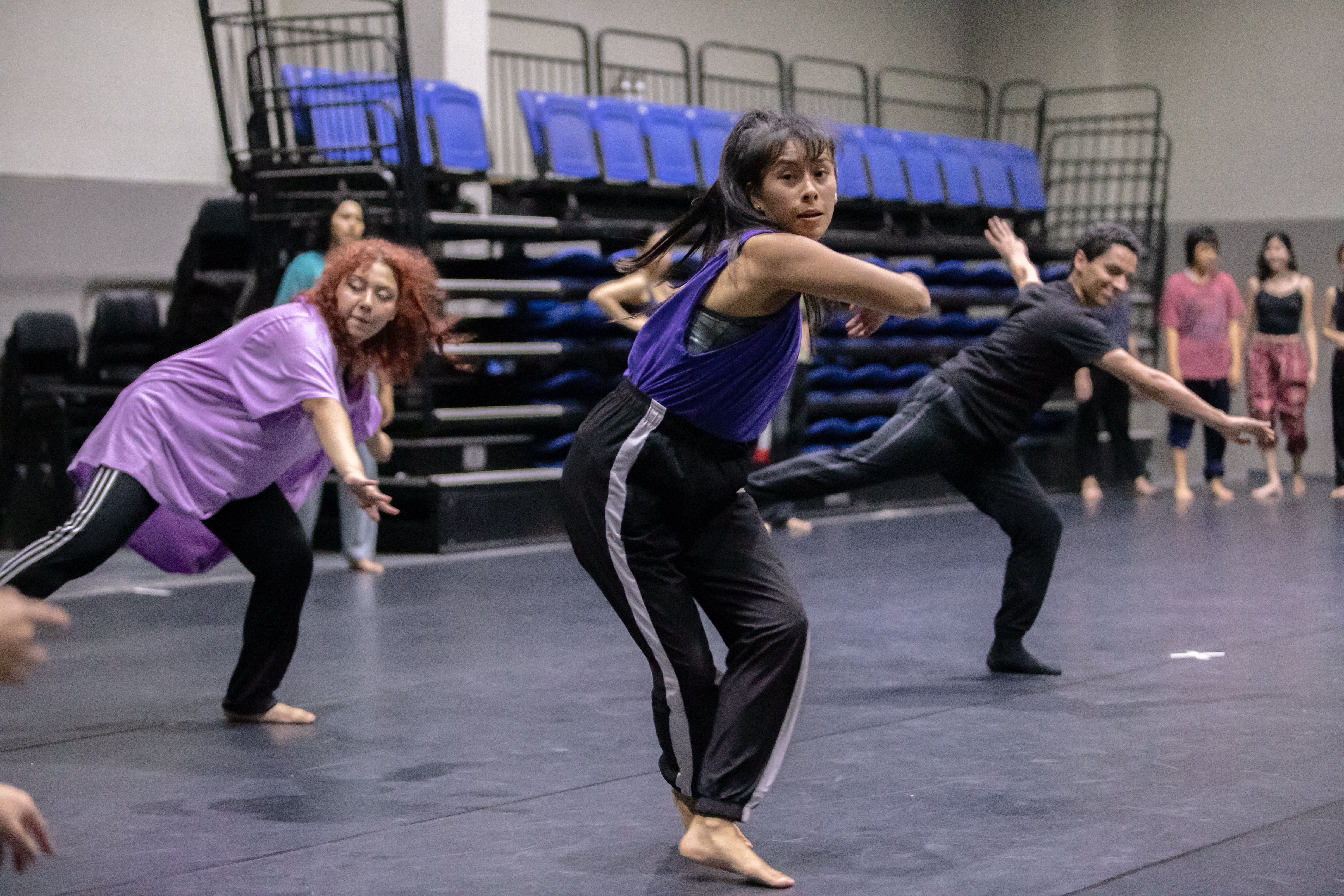 Fotografía que muestra en primer plano a una bailarina con ropa de entrenamiento realizando un movimiento de danza, otros dos bailarines repiten ...