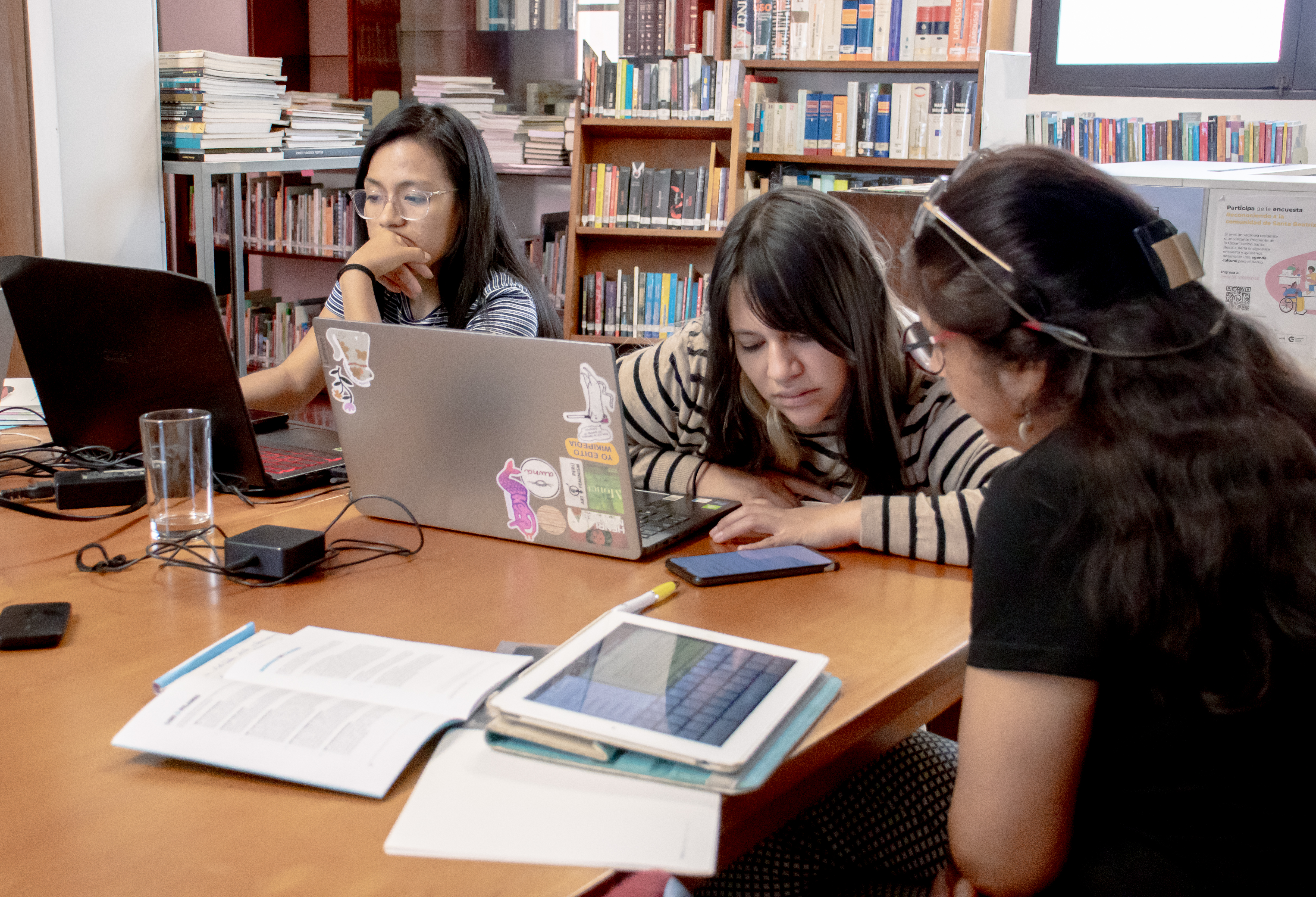 Foto que muestra en una bliblioteca a tres chicas trabajando frente a sus computadoras y a la vez intercambiando ideas.