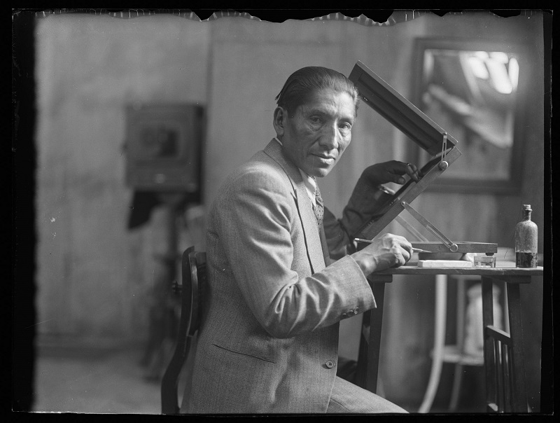 Fotografía en blanco y negro del fotógrafo Martín Chambi en su estudio, sentado ante una mesa de trabajo donde tiene un aparato de visualización de imágenes. Él mira a la cámara mientras sostiene en una mano un lápiz y con la otra acciona el aparato.