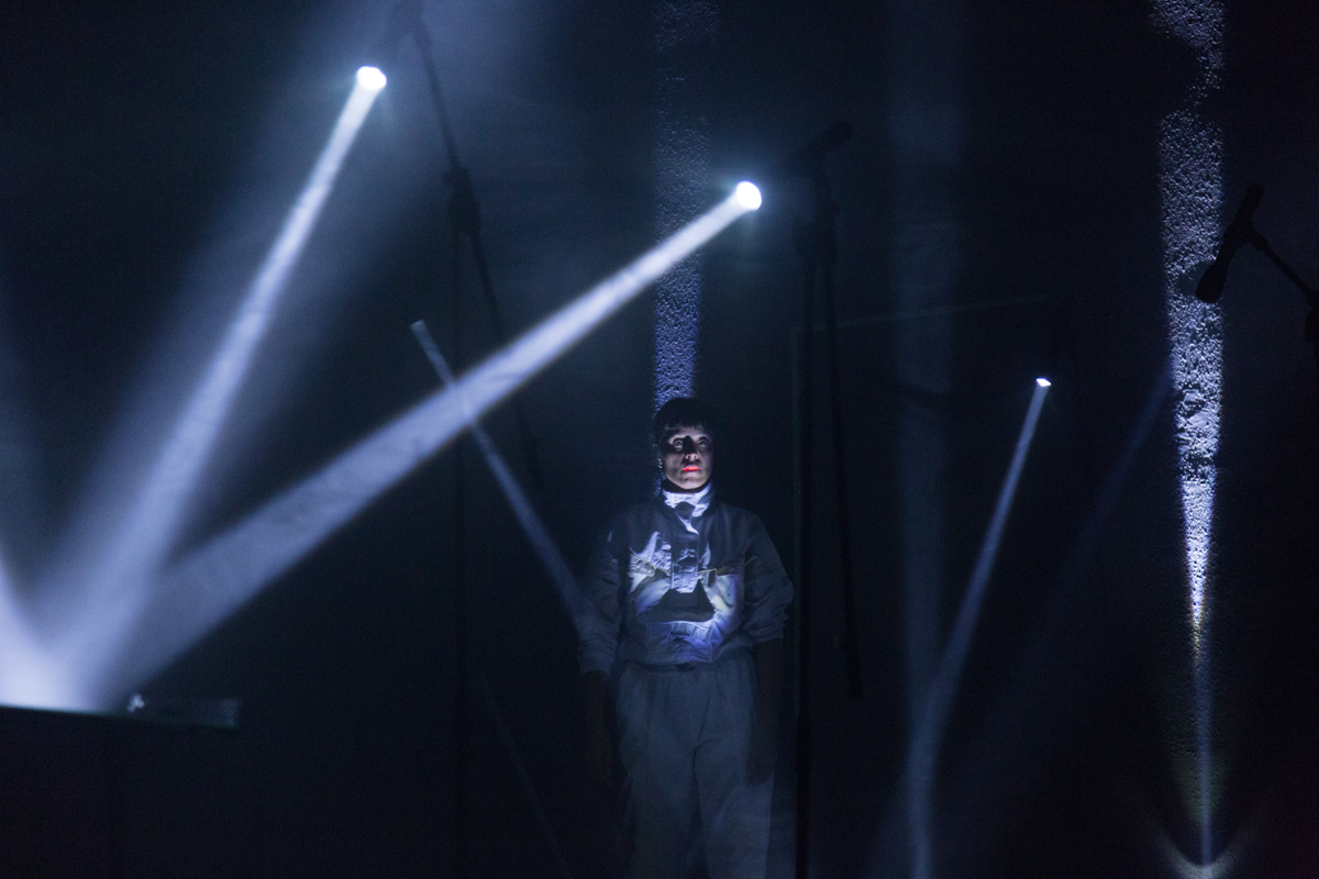 Foto de un espacio lleno de sombras y aces de luces que salen de varios focos. En medio de ese claroscuro hay una mujer parada en la que resaltan sus labios rojos y la ropa enteriza blanca que viste.