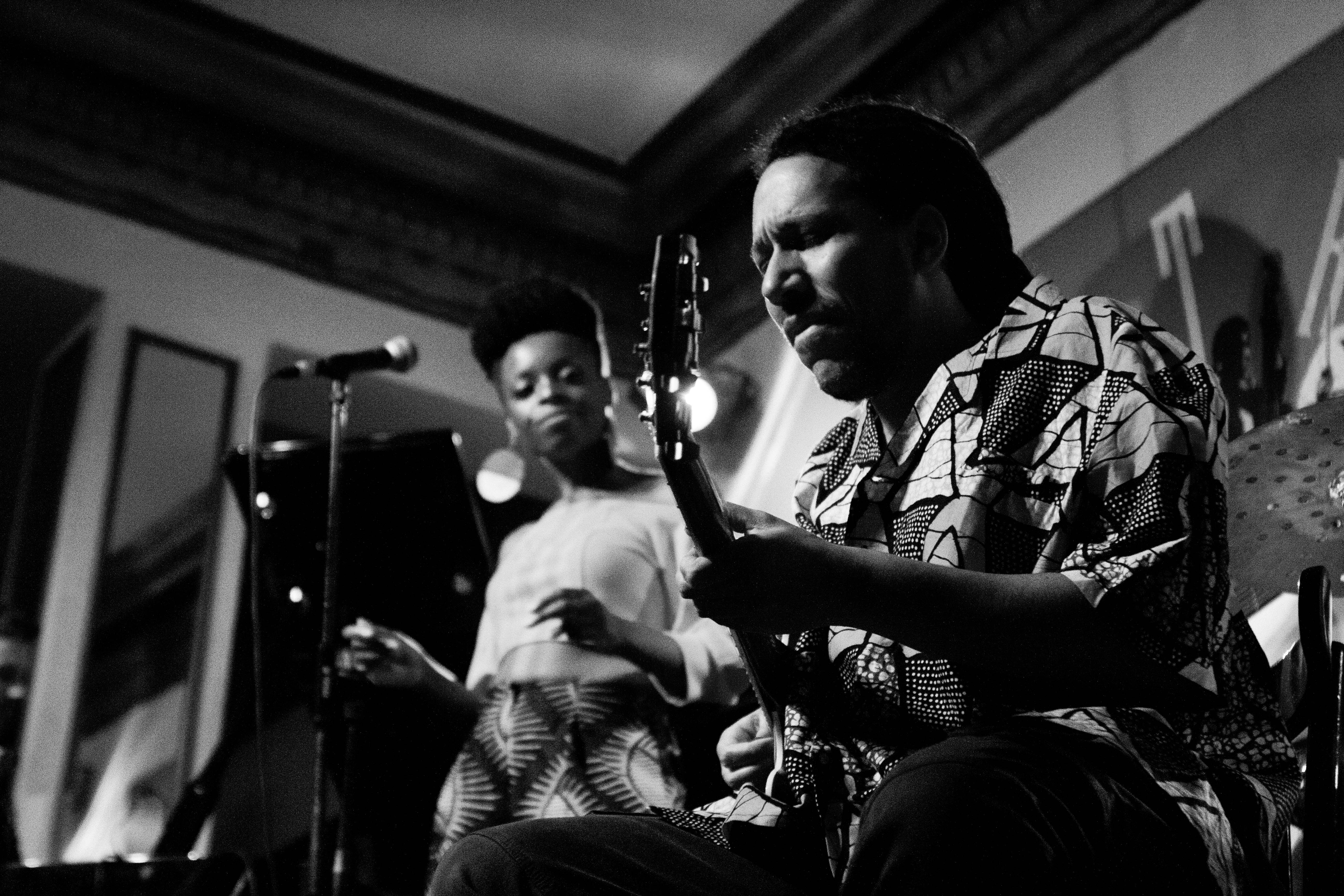 Foto en blanco y negro que muestra en primer plano a un guitarrista afrodescendiente que con los ojos cerrados interpreta una pieza en su guitarra. Está vestido con una camisa de temas africanos y lleva el pelo peinado en trenzas largas recogidas en la nuca. En segundo plano, se observa la imagen más desenfocada de una cantante a la que él acompaña.