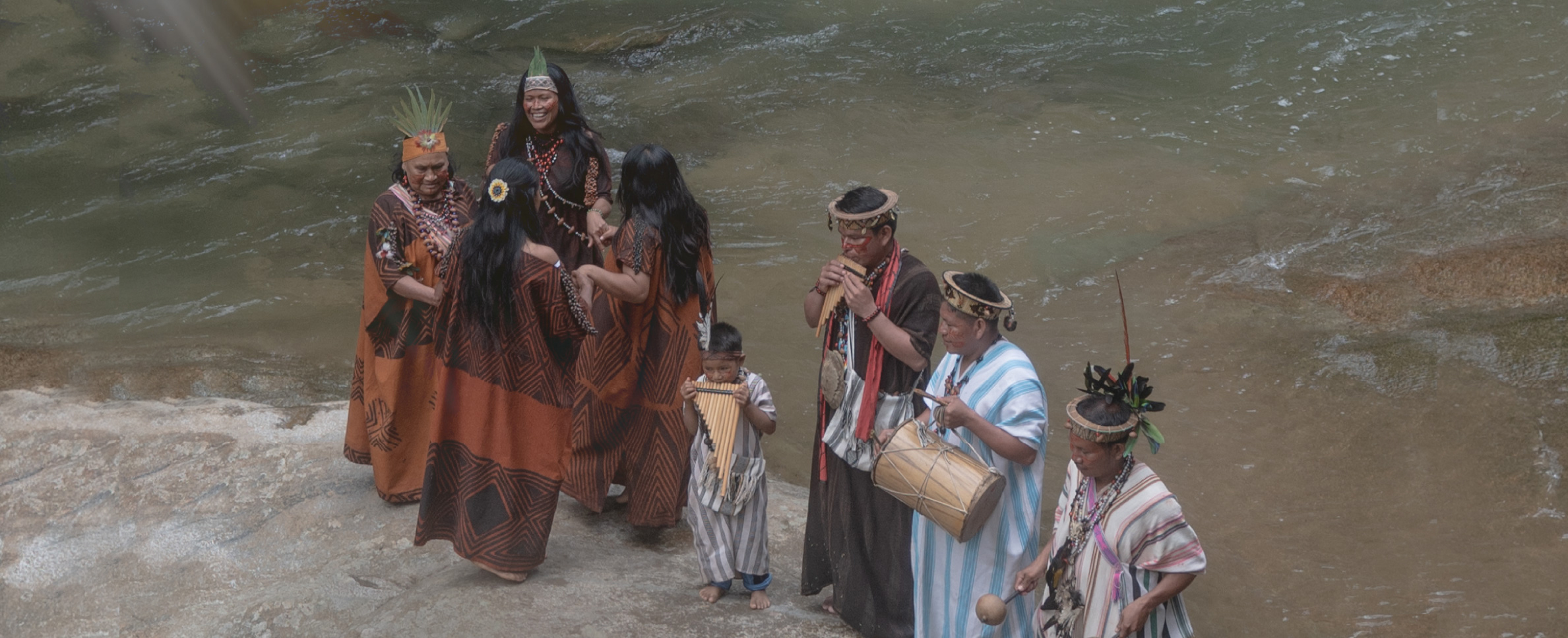 Foto que muestra, a la orilla de un río caudaloso, a un grupo de mujeres, hombres y un niño, ataviados con ropas y adornos propios de etnias amazónicas. Lad mujeres sonríen mientras hacen una ronda, y los hombres tocan instrumentos musicales como la zampoña, el tambor y las maracas.