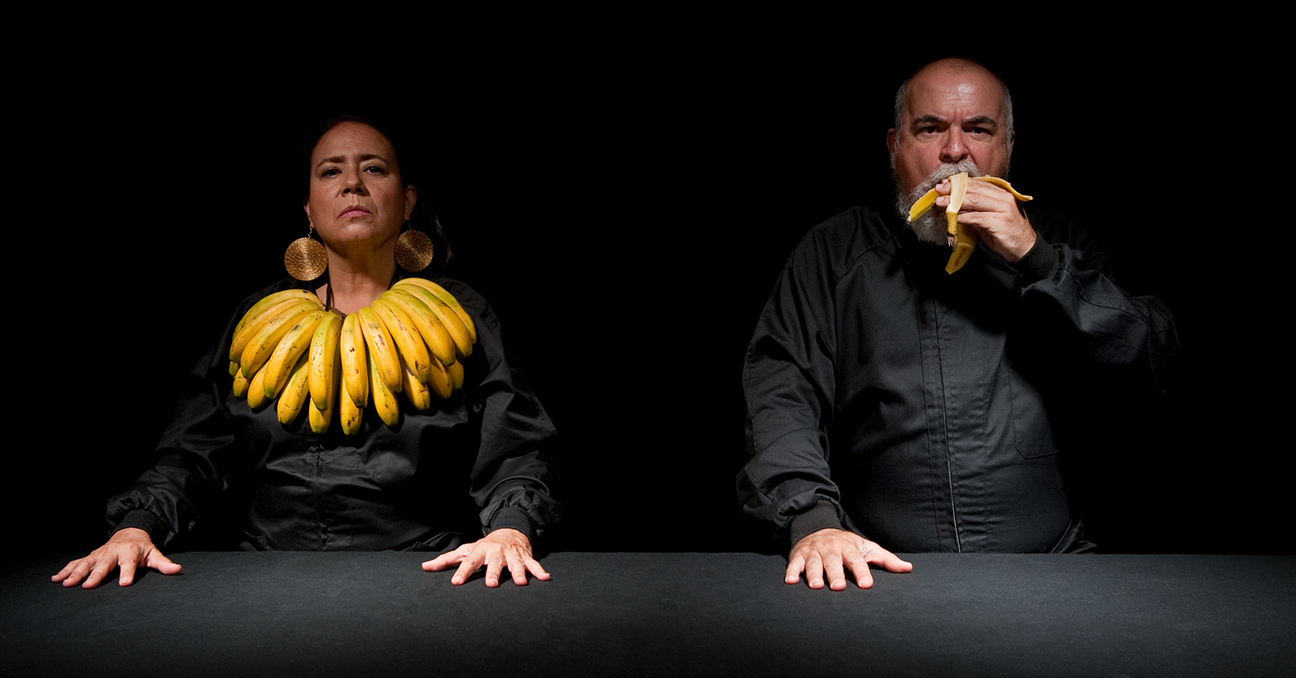 Una mujer con una ristra de bananos como collar y un hombre que se está comiendo un banano